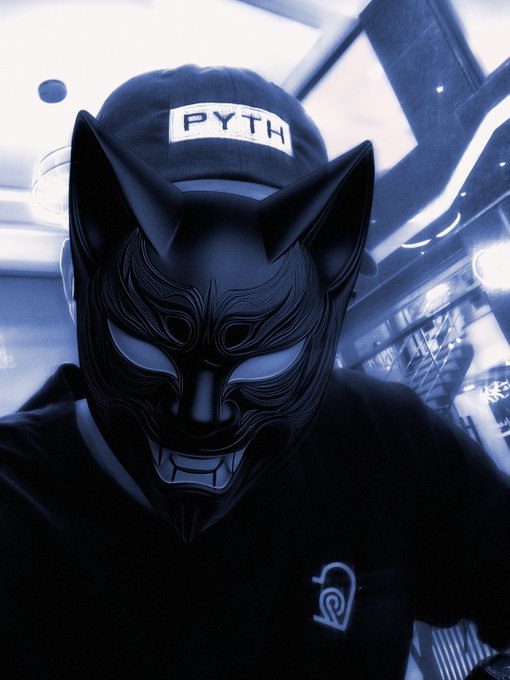 A person wearing a dark cap with a 'PYTH' patch and a black Kitsune-style demon mask, photographed in a blue-tinted setting.
