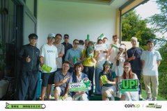 The image features a group of people posing for a photo, all holding up signs that say "coinup" or "coinup". The group consists of at least 13 individuals, with some standing closer together than others. They are gathered outdoors on a patio area, possibly in front of a building. In the background, there is a car visible, suggesting that this gathering might be taking place near a parking lot or a similar location.