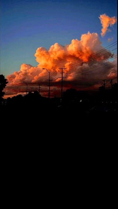 A scenic view of a vibrant sunset featuring orange, illuminated clouds against a darkening blue sky, silhouetted by power lines and streetlights over a dark foreground.
