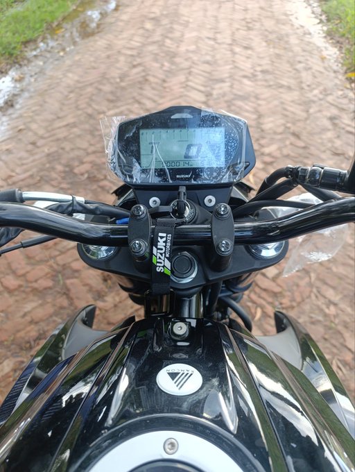 A newly purchased Suzuki motorcycle sits on a brick path, with the dashboard wrapped in protective plastic and the key in the ignition, reflecting the excitement of a "first bike" purchase made possible by Web3/crypto earnings. The visible "SUZUKI" branding on the handlebar clamp and the digital display showing "0 km/h" emphasize that this is a brand-new ride ready for its maiden journey.