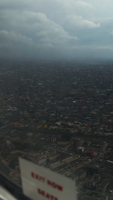 An aerial view of a densely populated cityscape is seen through a window with an "EXIT NOW ROWS SEATS" sign in the foreground. This image likely depicts Lagos, Nigeria, as suggested by the accompanying text stating "this una Lagos dey smell o," which can translate to "this Lagos is smelly."