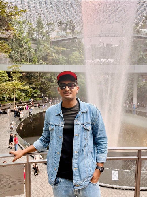 A man wearing a red cap, sunglasses, denim jacket, and black t-shirt posing at what looks like Jewel Changi Airport with the famous Rain Vortex waterfall in the background.