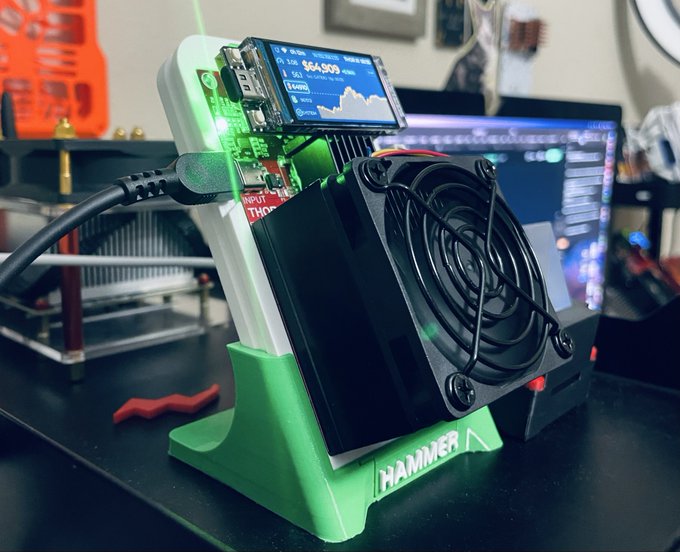 A compact cryptocurrency mining rig labeled 'HAMMER' with a green 3D-printed stand, featuring a small screen displaying Bitcoin data showing '$64,909' and active cooling fans.