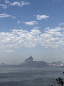 A coastal view of mountains rising from the ocean under a blue sky with scattered clouds, likely Rio de Janeiro.