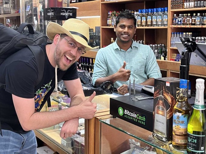 Two men smile and give thumbs up at a liquor store counter that displays a green 'Bitcoin Cash Accepted Here' sign next to various bottles of alcohol.