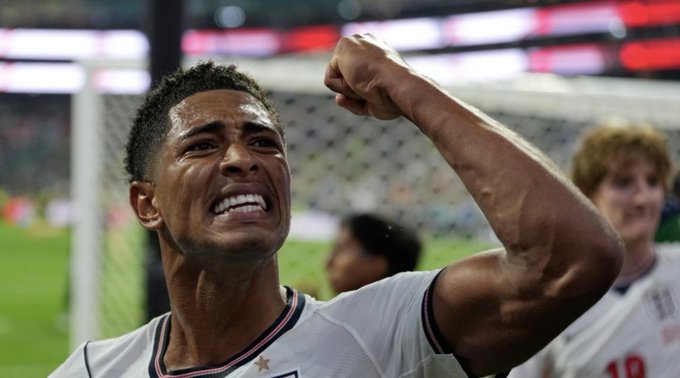 A close-up of a sweaty soccer player in a white jersey clenching his fist in an emotional celebration, with teammates visible in the background.