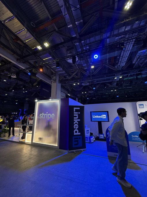 Convention floor view with illuminated booth displays for Stripe and LinkedIn, featuring a man in a blue suit walking past under industrial ceiling lighting.