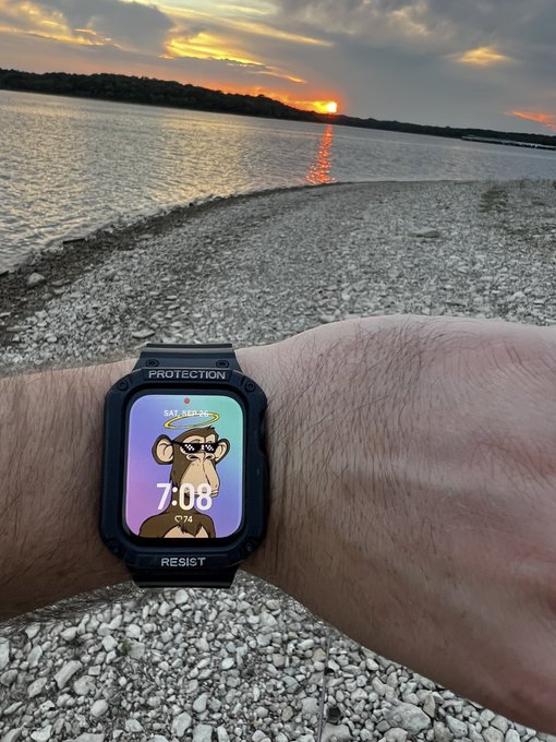 A wrist wearing a black smartwatch with a Bored Ape watch face displays '7:08' and 'SAT, SEP 20' with '°74' temperature, set against a sunset over a calm body of water with rocky shore.