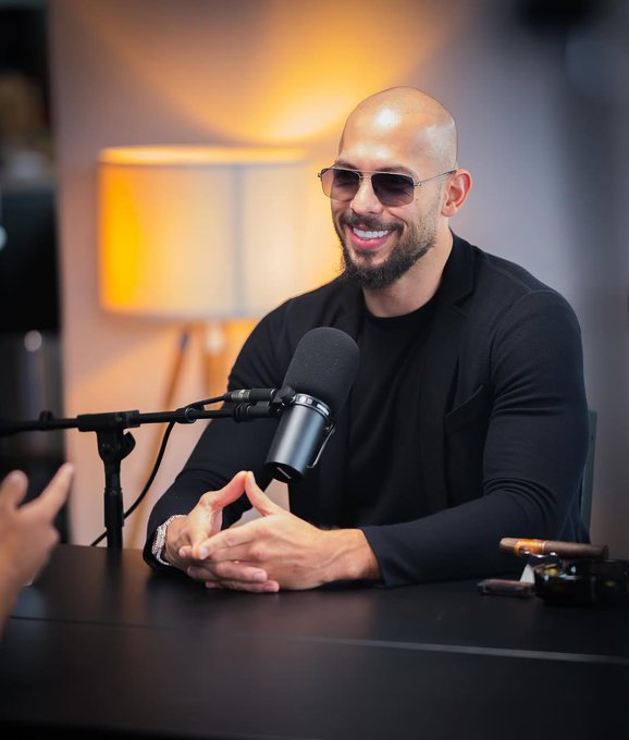 A bald man with a beard wearing sunglasses and a black jacket smiles while sitting at a podcast microphone in a studio setting.