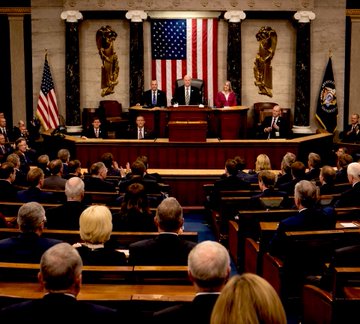 The image shows a group of people sitting in chairs, with some wearing suits and ties. They are gathered around a podium where a man is giving a speech or presentation. The room has an American flag displayed prominently above the podium. In the background, there are columns that suggest this could be a formal event or meeting. A post on social media indicates that Bitcoin and crypto clarity bills have been added to the Senate legislative calendar for voting.