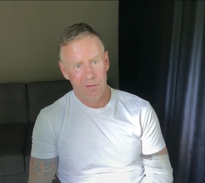A man with short blond hair and a tattoo on his arm, wearing a white t-shirt, sits on a couch with black curtains behind him, while his left arm appears wrapped in bandages. The context suggests the post jokingly reveals this image as the long-awaited appearance of Hodlonaut, tying into the surrounding joke about the BIP110 drama.