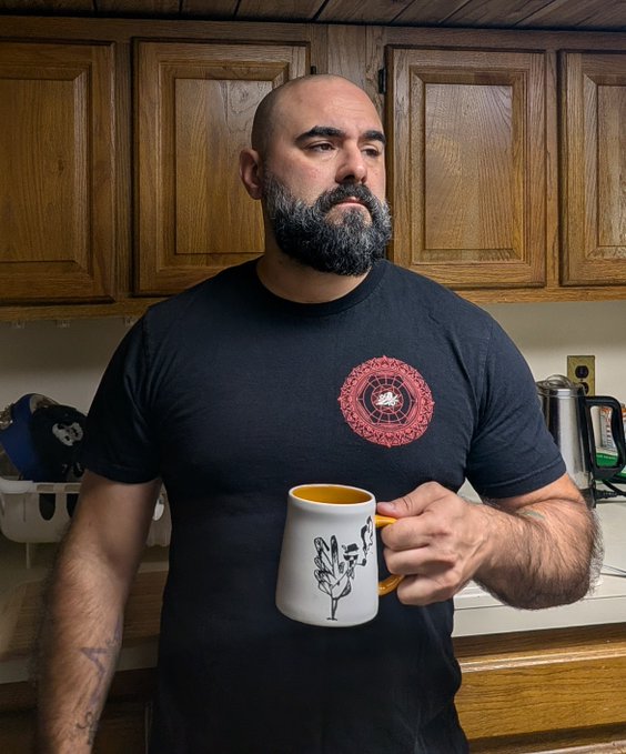 A bald man with a thick beard wearing a black t-shirt with a red circular mandala design, holding a white mug with a flower design in a kitchen.