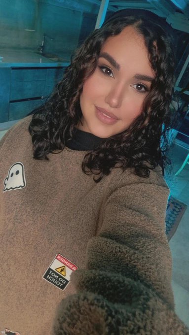 A selfie of a young woman with curly dark hair wearing a brown fleece sweater featuring a ghost patch and a 'DO NOT TOUCH' label.