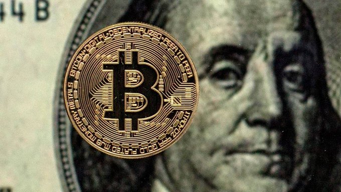A physical Bitcoin token rests on top of a US one hundred dollar bill featuring Benjamin Franklin's portrait.