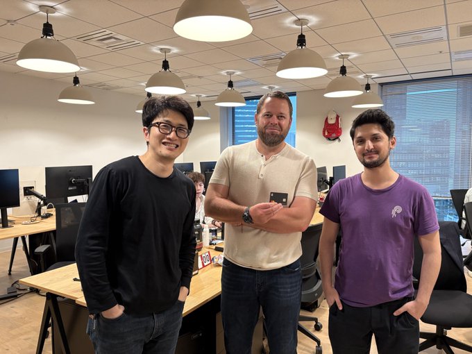 Three men pose together in a modern office setting with pendant lights and workstations, while a partially visible sign on the desk displays "BACK" — likely referencing Backpack Exchange, aligning with the post's promotion of the $BP token and its hardware wallet card shown by the central figure holding a small device.