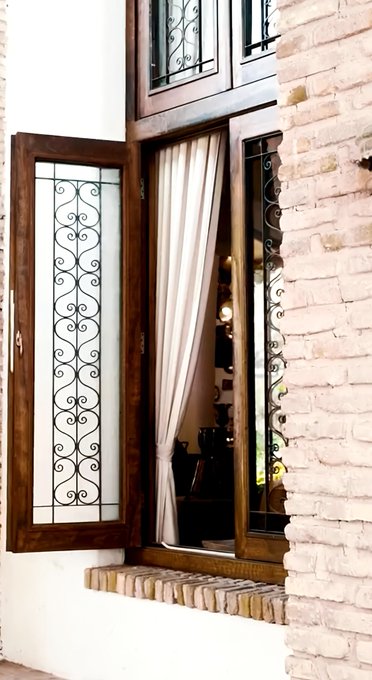 An open wooden casement window with decorative wrought iron detailing reveals a cream-colored curtain, set into a whitewashed brick wall.