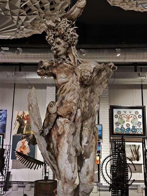 A large, weathered sculpture of a humanoid figure with an elaborate horned headdress stands in a gallery space surrounded by framed artworks and industrial decorative elements.