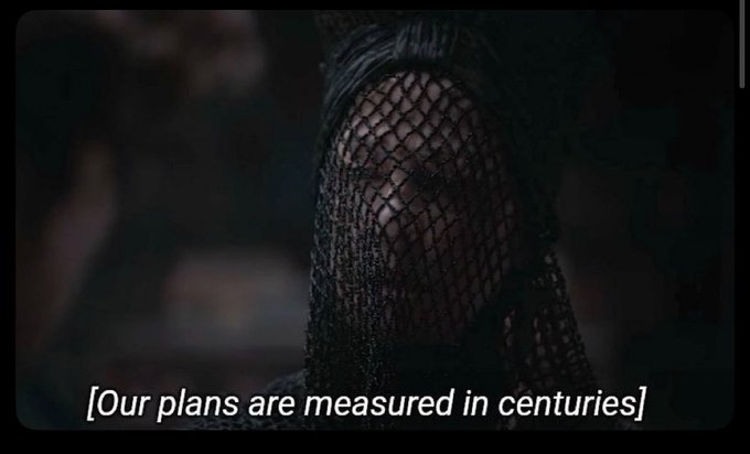 A close-up of a person wearing a dark net veil over their face, with the subtitle '[Our plans are measured in centuries]' visible at the bottom.