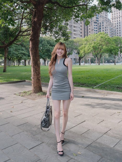 A woman with long auburn hair poses in a grey sleeveless mini dress and black heels on a park pathway, holding a black handbag with trees and apartment buildings behind her.