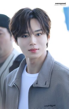 A close-up photograph of a young man with brown hair wearing a grey jacket over a white t-shirt, with 'overexposed' watermarked in the corner.