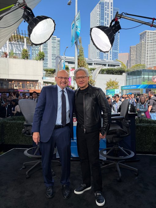 Two men, one in a navy suit and the other in a black leather jacket, pose together at an outdoor broadcast setup with CNBC and Salesforce signage visible, alongside an Omniverse XR Stage display, at what appears to be a tech industry event in downtown San Francisco. The caption "If Nvidia starts dumping you already know why…" appears to humorously reference the billionaire tech mogul on the right (Jensen Huang, Nvidia's CEO) being the reason behind any market volatility involving his company.