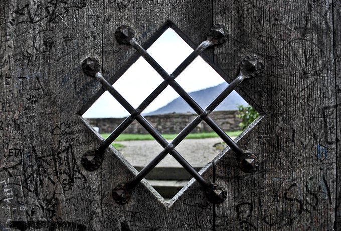 A diamond-shaped window with a black metal lattice grate set in a weathered, graffiti-covered wooden door, revealing a view of grass, stone walls, and distant mountains outside.