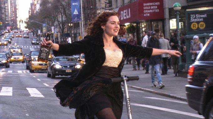 A woman in a black dress and lace top joyfully rides a scooter down a busy New York City street, holding a bottle of Jack Daniel's whiskey, near the 23 Street Station.