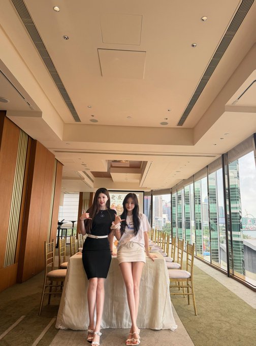 Two young women in stylish outfits (a black dress and a white outfit) pose together at a long dining table in an upscale restaurant with floor-to-ceiling windows overlooking a city skyline.