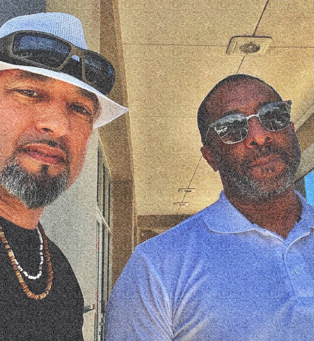 Two men pose for a selfie; the man on the left wears a white hat, sunglasses, and a beaded necklace, while the man on the right wears a blue shirt and sunglasses.