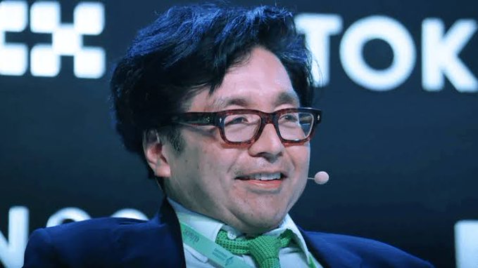 A bespectacled man wearing a green tie and headset microphone smiles while speaking on stage, with partial "TOKYO" text and a "Citi" logo visible in the background — likely Tom Lee, referenced in the post discussing Bitmine's $74 million Ethereum accumulation and broader crypto market positioning.