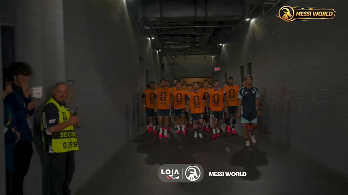The image shows a group of people walking down an empty hallway, wearing orange jerseys with numbers on them. There are at least 13 people visible in the scene, some closer together while others are more spread out. The hallway appears to be long and narrow, possibly leading towards a specific destination or event. In the background, there is a sign that reads "MESSY WORLD", which might indicate the location of an event or gathering taking place within this building.