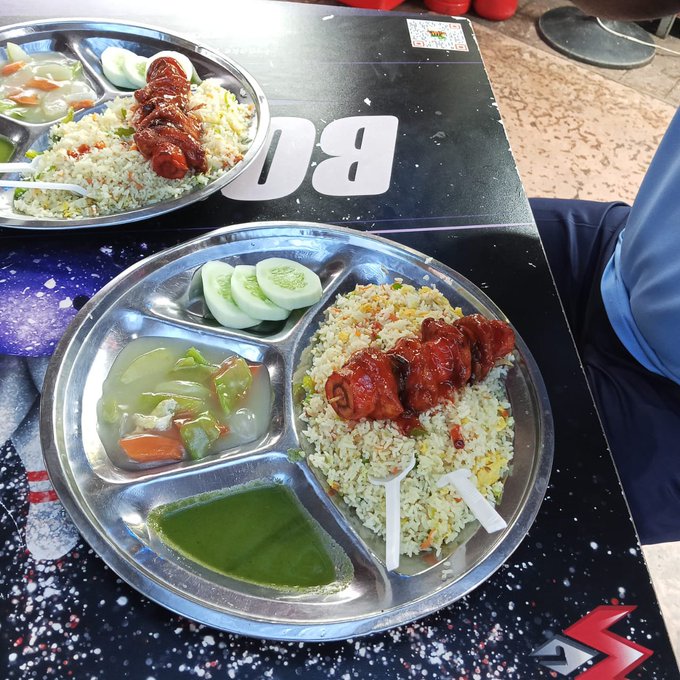 Two compartmentalized metal plates containing rice, red-marinated meat skewers, sliced cucumbers, and vegetable curry, served on a table with a space-themed design.