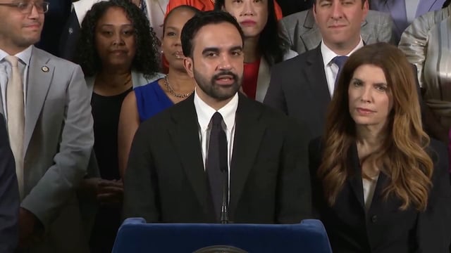 A man in a dark suit speaking at a podium with a presidential seal, surrounded by a crowd of onlookers including men in suits and women in formal attire.