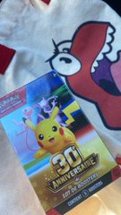 A Pokémon trading card featuring Pikachu lies on a white shirt with a red character design, with the text "30th ANNIVERSARY" visible on the card.