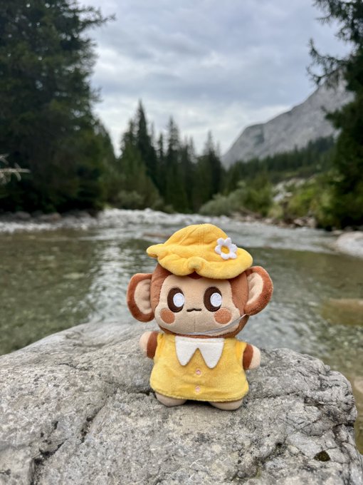 A plush monkey character wearing a yellow dress and a small flower sits on a rock beside a mountain stream, fitting the cheerful "Gm folks, have a happy Chuesday" post celebrating !CHIMP.