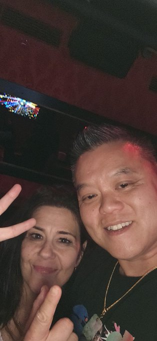 A casual selfie of a smiling man and woman posing together indoors with a peace sign, with colorful lights visible in the background.