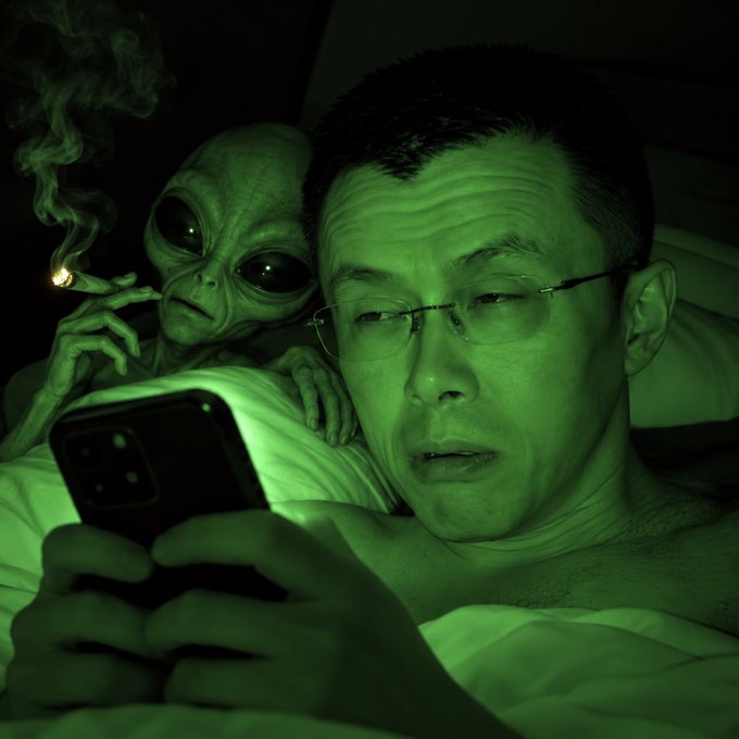 A night-vision style, green-tinted photo of a man wearing glasses looking at a smartphone while lying in bed, with a grey alien prop over his shoulder holding a smoking cigarette.