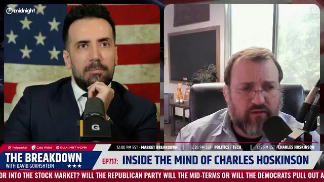 The image shows two men sitting at a table with microphones in front of them. One man has a beard and appears to be wearing a suit, while the other man has glasses and seems to be dressed more casually. They are both talking into their microphones during an interview or discussion on television. The background features an American flag, which is prominently displayed above the two men's heads.
