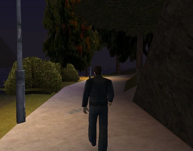 A video game character in dark clothing walks down a paved path at night, flanked by bushes, trees, and a streetlight.