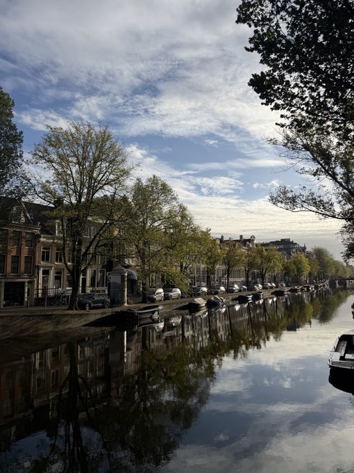 A serene canal lined with parked cars and traditional Dutch buildings reflects a dramatic, cloud-filled sky, capturing a peaceful morning scene in what appears to be Amsterdam as a stroll to the office begins.