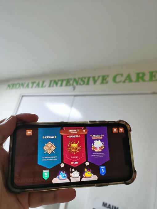 The image shows a person holding up their cell phone displaying a game screen with three different levels, each featuring a character or item from the game. The person's hand is positioned over the top of the phone while they are looking at the screen. The background features a green sign that reads "Neonatal Intensive Care". This suggests that the person might be waiting for their baby to arrive and is playing a game on their cell phone as a form of entertainment or distraction during this time.