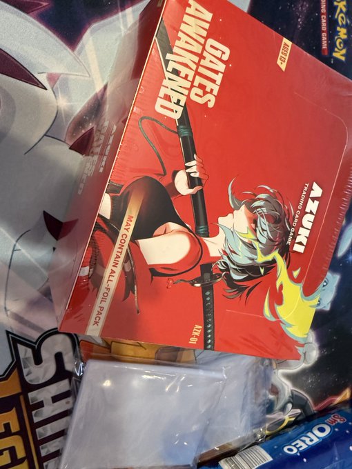 The image shows an open red box containing a game, with a cartoon character on the front of the box. The box has been opened, revealing its contents. There are several items inside the box, including a game, a toy car, and some other objects. A post on social media is visible in the background that reads "Opening a 2nd @AzukiTCG box - wish me luck".