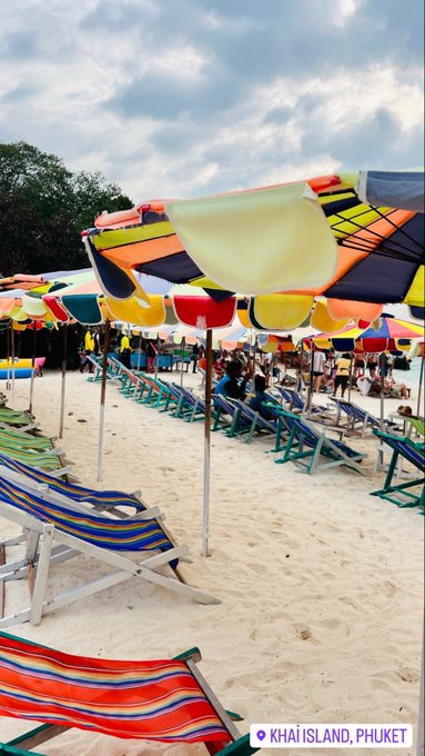Colorful striped umbrellas and lounge chairs line the sandy beach at Khai Island, Phuket, with people gathered under the bright canopies against a partly cloudy sky. The vibrant, relaxed setting captures the "Gm Gm" (good morning) vibe perfectly, offering a cheerful tropical welcome to the day.
