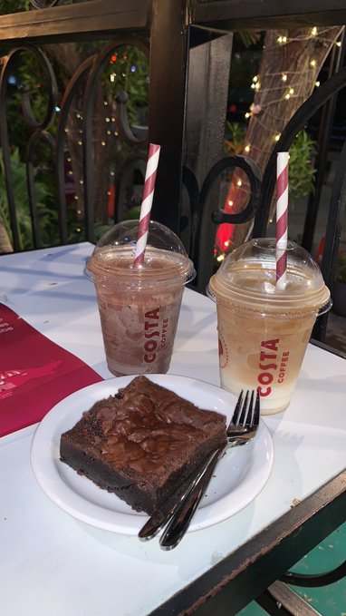 A photograph of two iced Costa Coffee drinks in plastic cups with red-and-white striped straws served on a white table alongside a plate with a chocolate brownie and a fork.