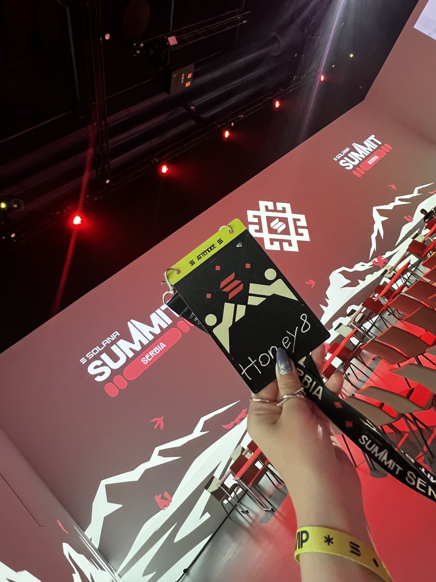 A first-person view of a hand holding up a black VIP lanyard with a 'Honey 8' pass at an event; the red backdrop reads 'SOLANA SUMMIT SERBIA'.
