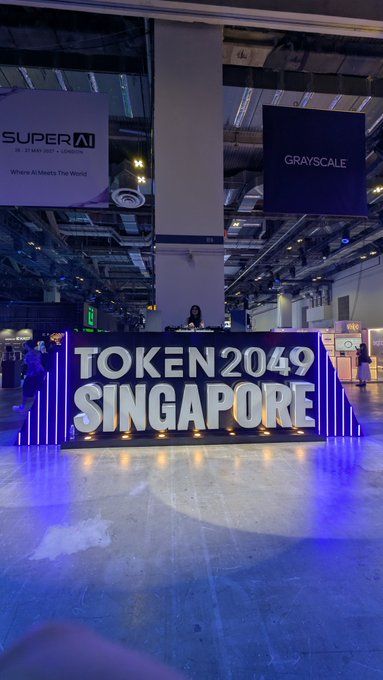 A large illuminated "TOKEN 2049 SINGAPORE" sign stands in a convention hall, with banners for "SUPER AI" and "GRAYSCALE" visible above, capturing the atmosphere of the crypto conference the post references.