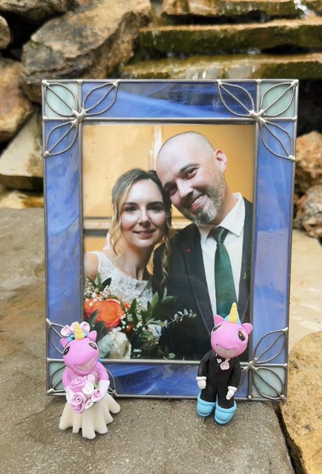 A stained-glass-style framed wedding photo of a smiling couple is displayed on stone, accompanied by two handmade clay unicorn figurines—a pink bride unicorn in a dress and a dark-suited groom unicorn—both wearing unicorn horns, with the post noting the thoughtful gifts were sent from France by @CharleyMakes.