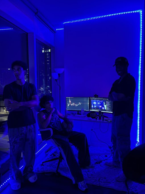 Three young people pose in a dim room bathed in blue LED light, with two computer monitors displaying a stock trading chart (including a visible candle chart on the left) and what appears to be a livestream feed on the right, tying into the "Live on Robinhood" crypto/stock trading stream being broadcast at doink.live/c/0x65ec40f99c.