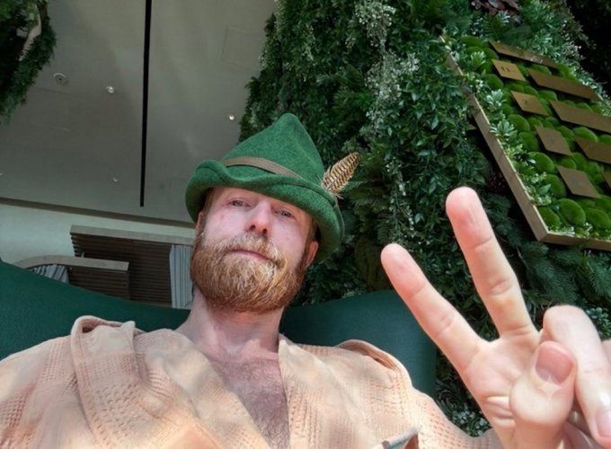 A bearded man wearing a green felt hat with a feather and a beige knit shirt takes a selfie, making a peace sign with his fingers.