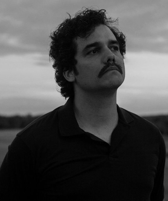 A black-and-white portrait of a man with curly dark hair and a thick mustache, wearing a dark polo shirt, gazing into the distance with a cloudy sky behind him.